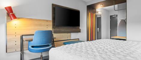 Una habitación de hotel moderna con una cama, un escritorio con silla, un televisor montado en la pared y un baño con espejo y toallero.