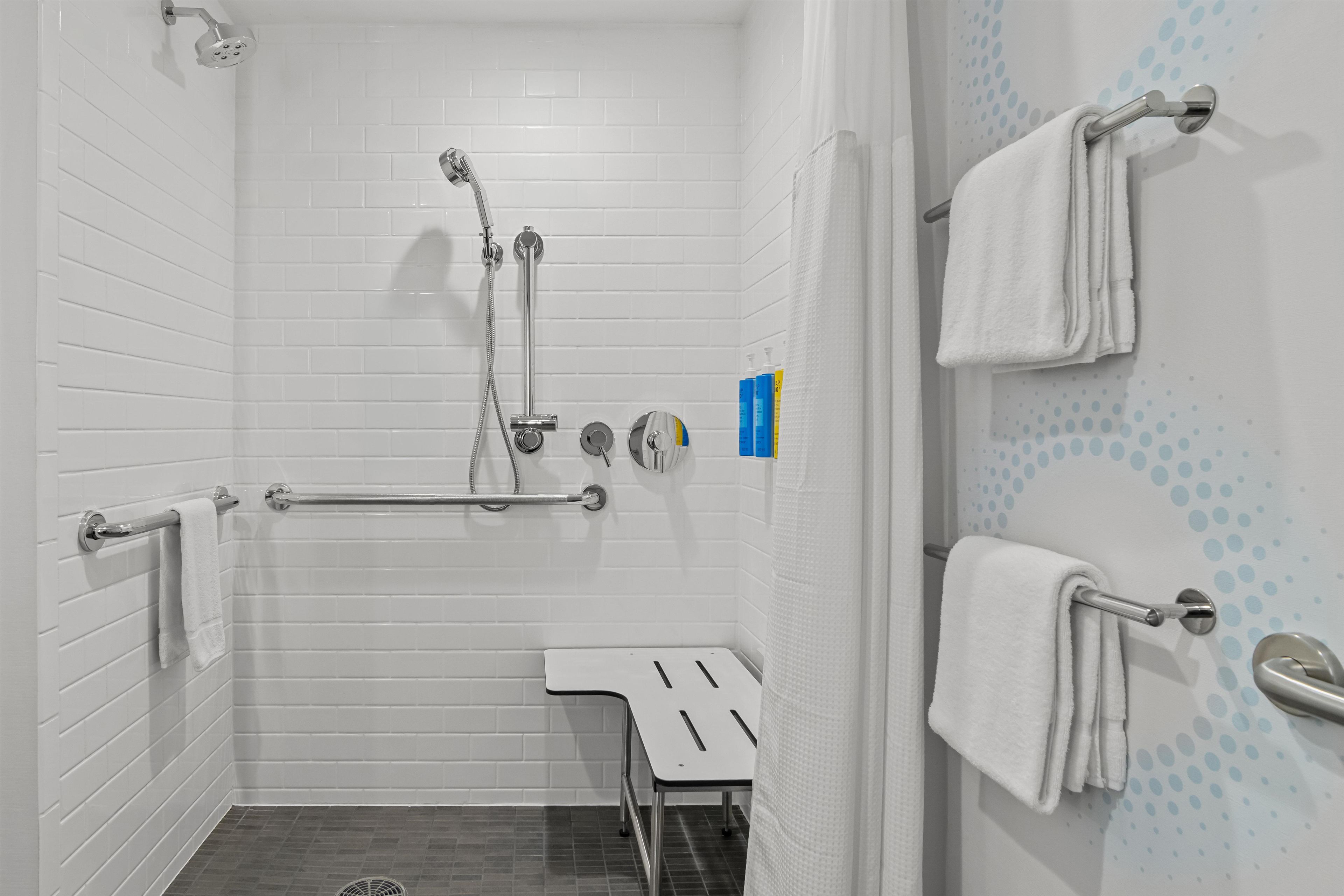 A bathroom with white tiled walls, a shower with a handheld showerhead, a grab bar, a towel rack, and a small bench.