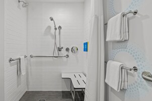 Un baño con paredes de azulejos blancos, una ducha con cabezal de ducha manual, un asidero, un perchero para toallas y un pequeño banco.