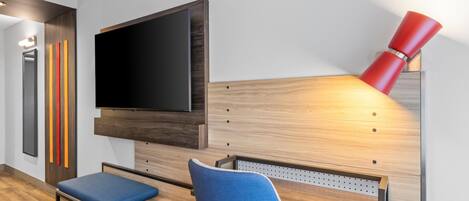 Ein modernes Hotelzimmer mit einem wandmontierten Flachbildfernseher, einer roten Stehlampe und einer blauen Polsterbank.