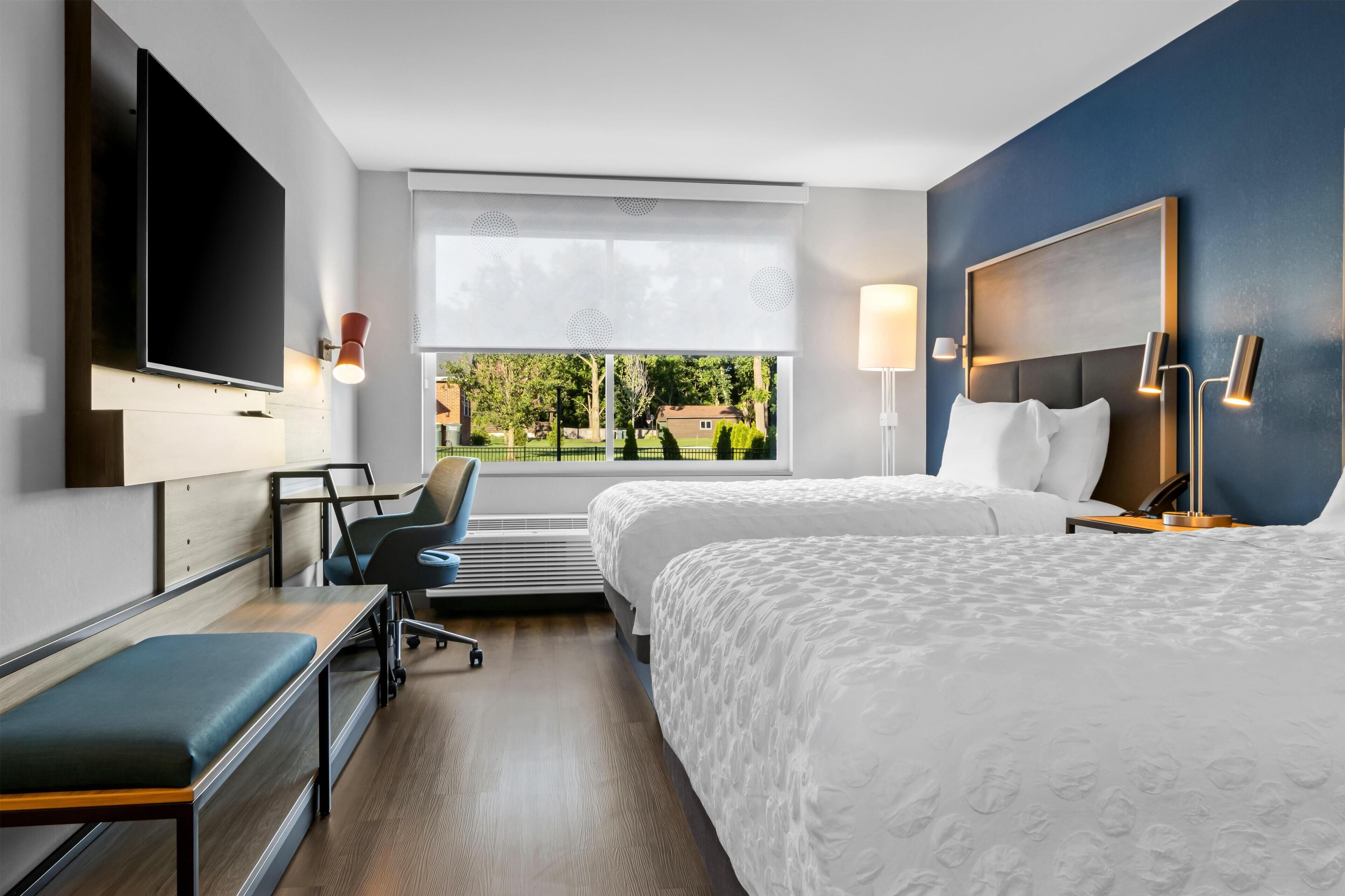 Une chambre d’hôtel avec un grand lit, un bureau avec une chaise, un téléviseur fixé au mur et une fenêtre donnant sur un paysage verdoyant.