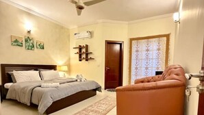 Un dormitorio con cama, ventilador de techo, mesita de noche, lámpara, alfombra, puerta y ventana con cortinas.