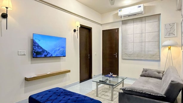 Un soggiorno moderno con un pouf blu, un divano grigio, un tavolino da caffè in vetro e una TV a schermo piatto montata a parete.