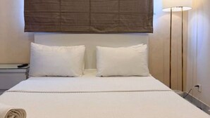 Camera d'albergo con un letto grande, due cuscini bianchi, una testiera, un comodino, una lampada e un asciugamano arrotolato sul letto.