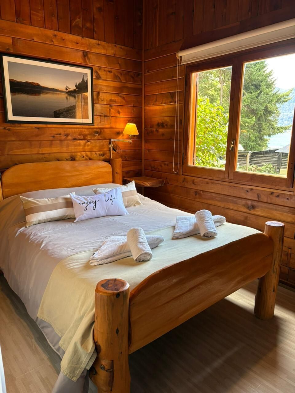 Quarto de cabana de madeira com cama, almofadas, mesa de cabeceira, abajur e janela.