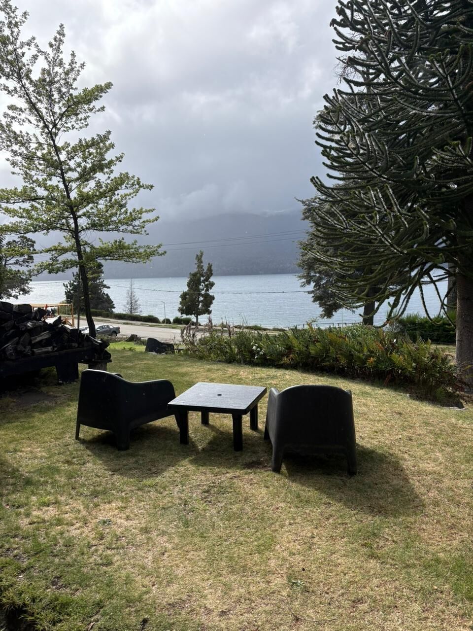 An outdoor seating area with a table and chairs, a pile of firewood, and a view of a body of water and mountains in the background.