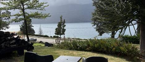 An outdoor seating area with a table and chairs, a pile of firewood, and a view of a body of water and mountains in the background.