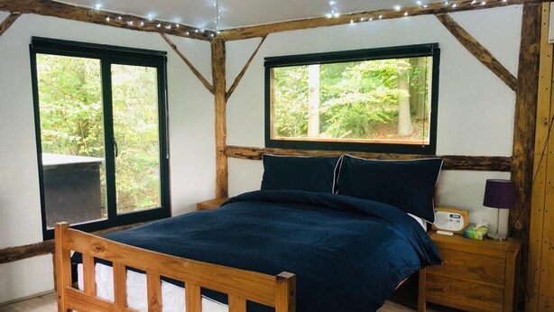 A wooden bed in a room with large windows, a bedside table, and a lamp. - Magical Treehouse/Cabin hidden in Ancient Woodland (Farnham)