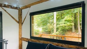 A room with a large window offering a view of a forested area. There is a wooden beam structure above the window, and a bed with dark bedding is visible. - Magical Treehouse/Cabin hidden in Ancient Woodland (Farnham)