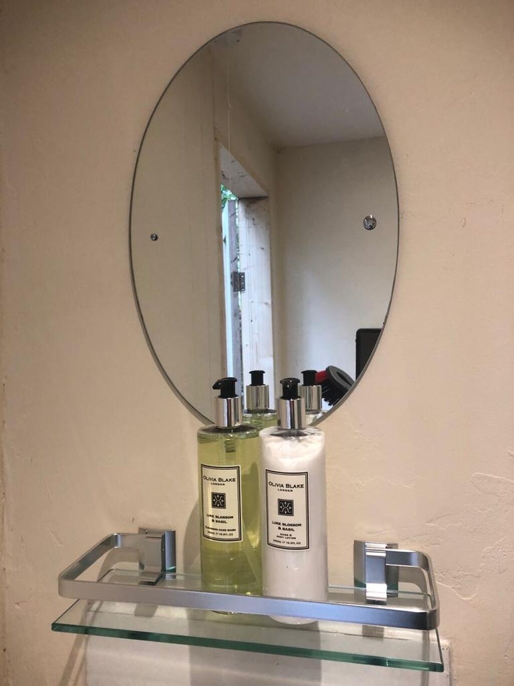 Een badkamer met een ronde spiegel, een glazen plankje en twee flessen Olivia Blake London body wash.