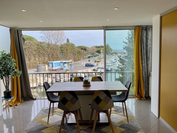 Un espace de repas moderne, doté d’une grande table et de chaises, offrant une vue sur un port de plaisance et comprenant un balcon avec des rideaux jaunes.