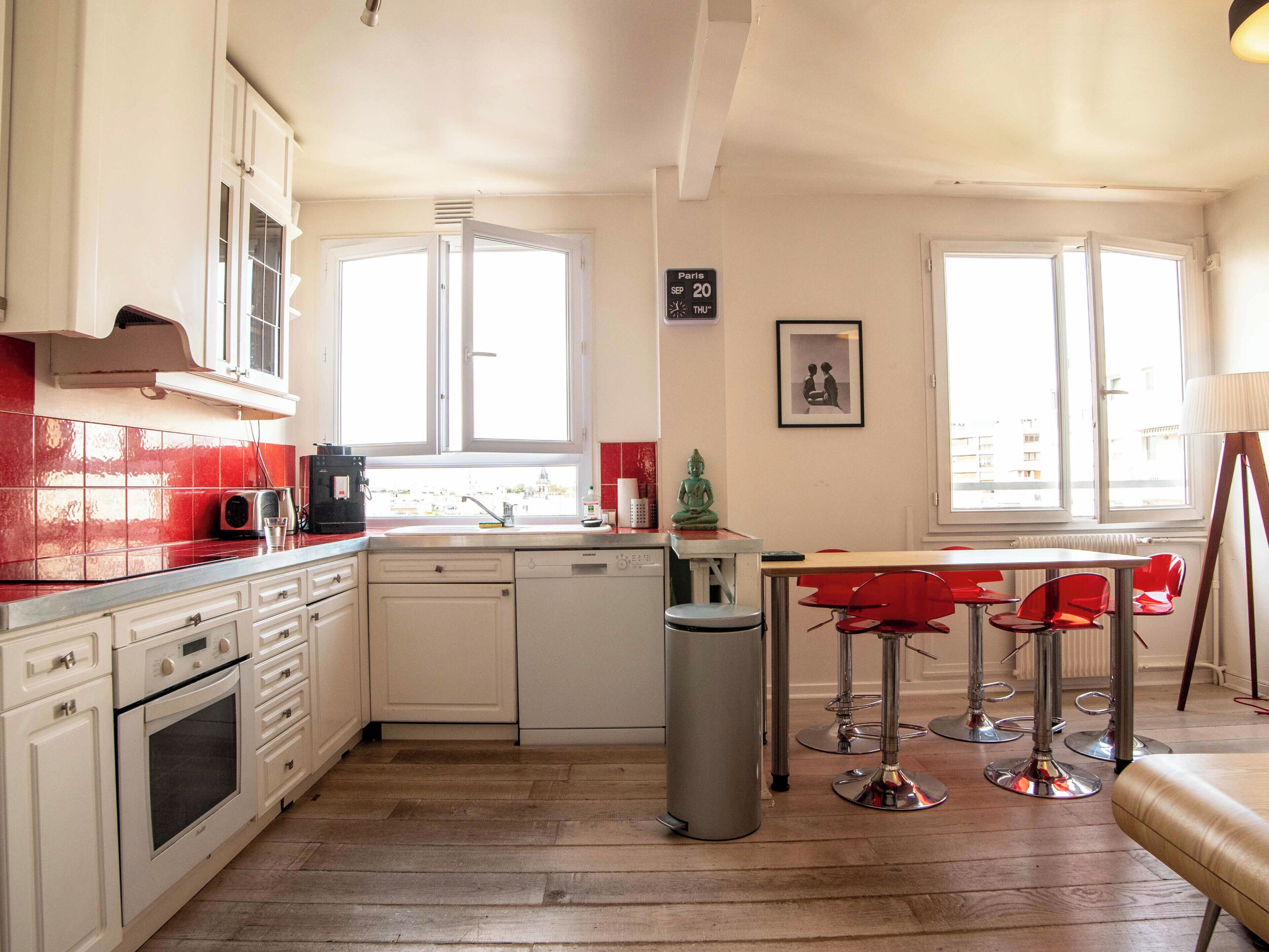 A kitchen with white cabinets, a red-tiled backsplash, a dishwasher, and a window overlooking a cityscape.