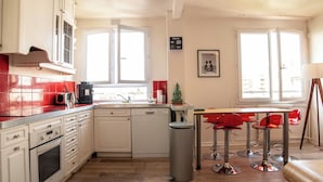 A kitchen with white cabinets, a red-tiled backsplash, a dishwasher, and a window overlooking a cityscape. - Spacious apartment near Père Lachaise (Paris)
