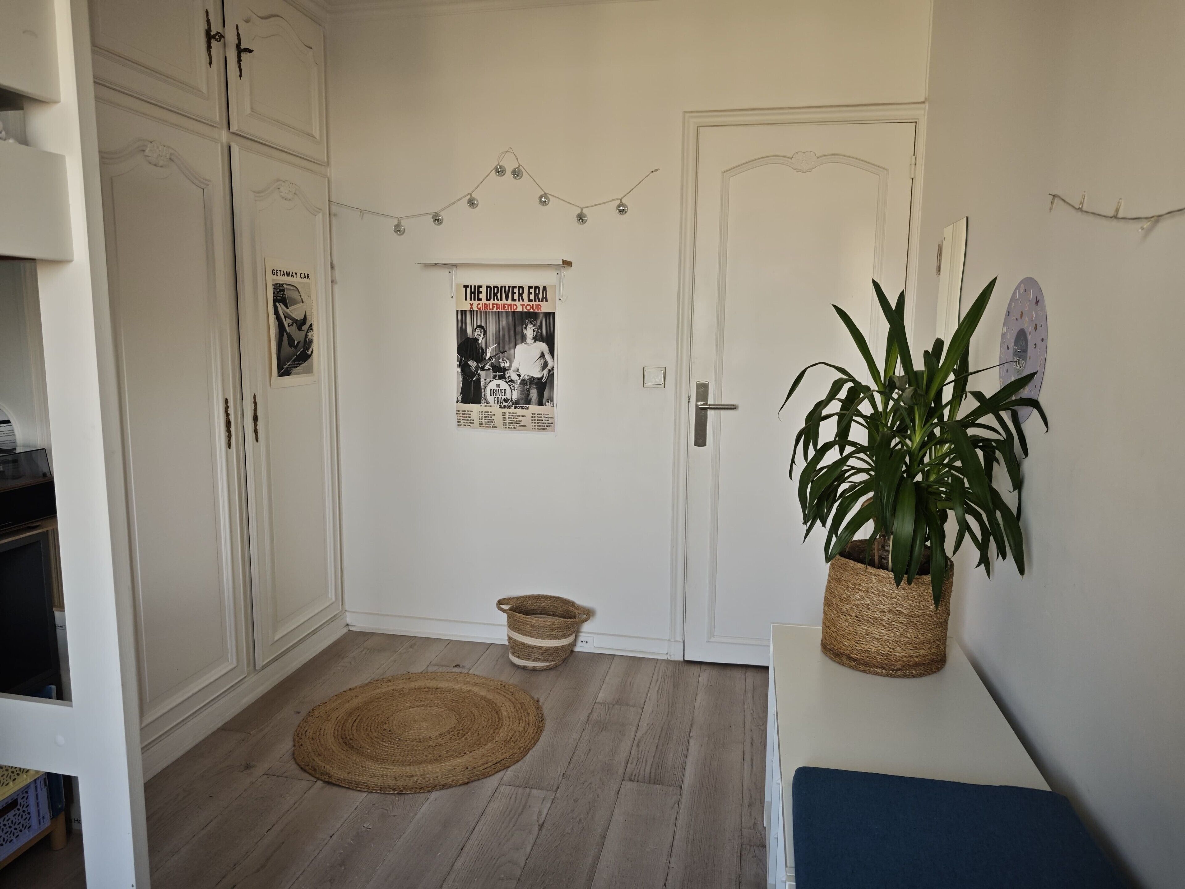 A hallway with a white door, a poster on the wall, a plant on a table, and a woven mat on the floor.