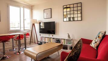 A modern living room with a red sofa, a wooden bench, a TV, a bookshelf, and a large window.