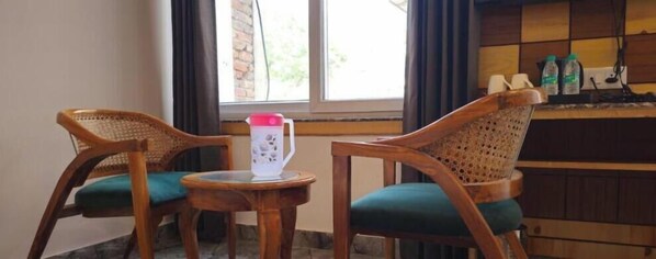 Two wicker chairs with teal cushions, a small round table with a white pitcher, and a window with curtains. - Hotel Dev (Rishikesh)