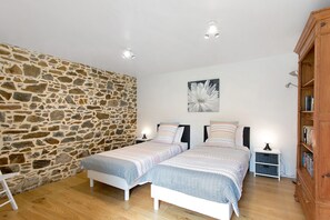 Un dormitorio con dos camas, una pared de piedra, suelo de madera, una mesita de noche, una lámpara y una estantería.