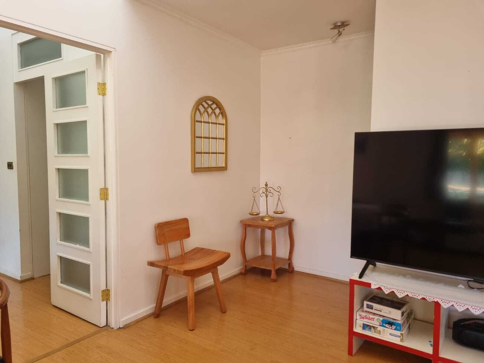 A living room with a wooden floor, a TV, a small table with a scale, and a window with a decorative frame.