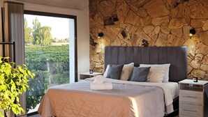 A bedroom with a stone wall, a bed with a quilted cover, two bedside tables, and a view of a vineyard through a large window. - Rincon Del Malbec (Luján de Cuyo)