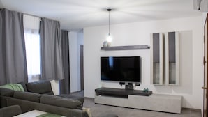 En moderne stue med et stort fladskærms-tv monteret på væggen, en grå sofa, et spisebord med stole og en pendel.