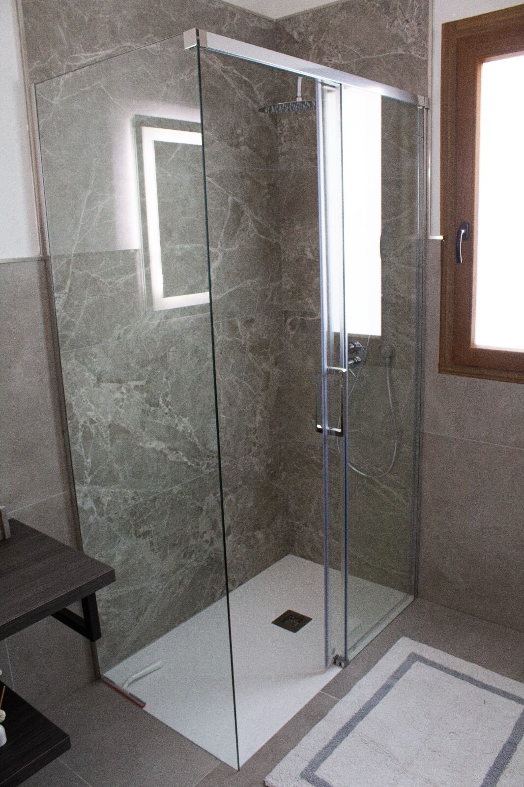 A glass-enclosed shower stall with a door, a wooden-framed window, and a small shelf with toiletries.