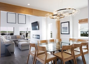 A modern living room with a glass dining table, wooden chairs, a sofa, and a TV mounted on the wall. - Steps to Gulf Beach & Private Lagoon + Amenities (Gulf Shores)