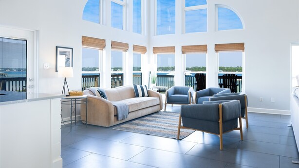 A spacious living area with a high ceiling, large windows, a sofa, chairs, and a view of the water.