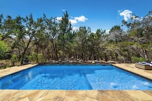 A rectangular swimming pool with clear blue water, surrounded by a stone deck and a natural landscape with trees and rocks.