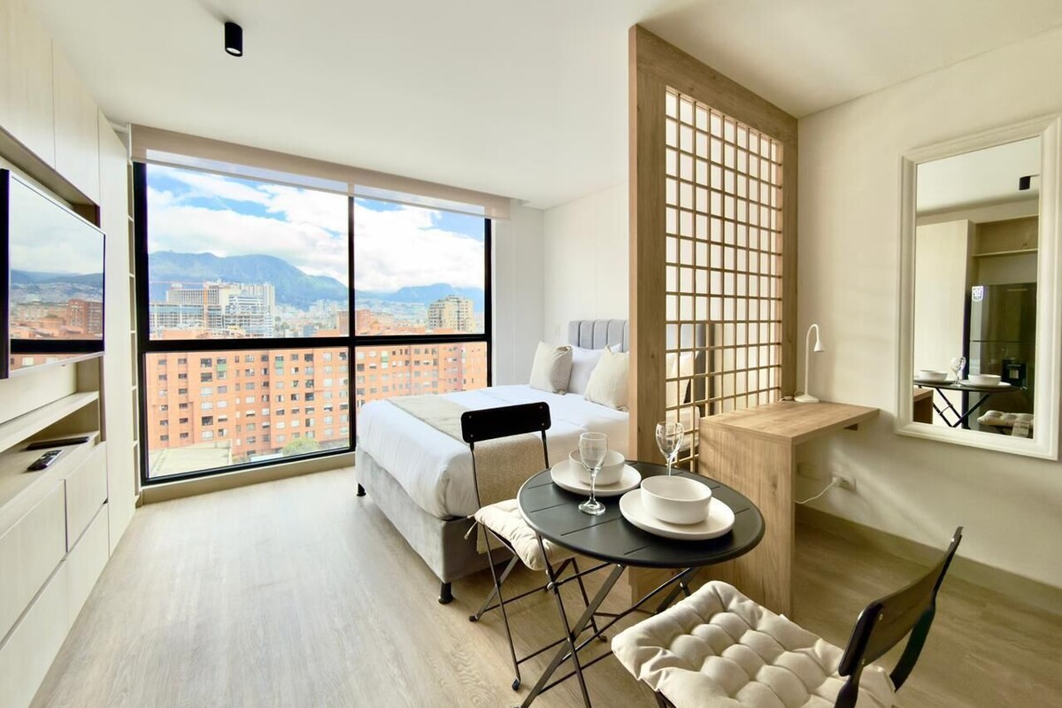 A modern hotel room with a large window, a bed, a dining table with chairs, and a small kitchen area.