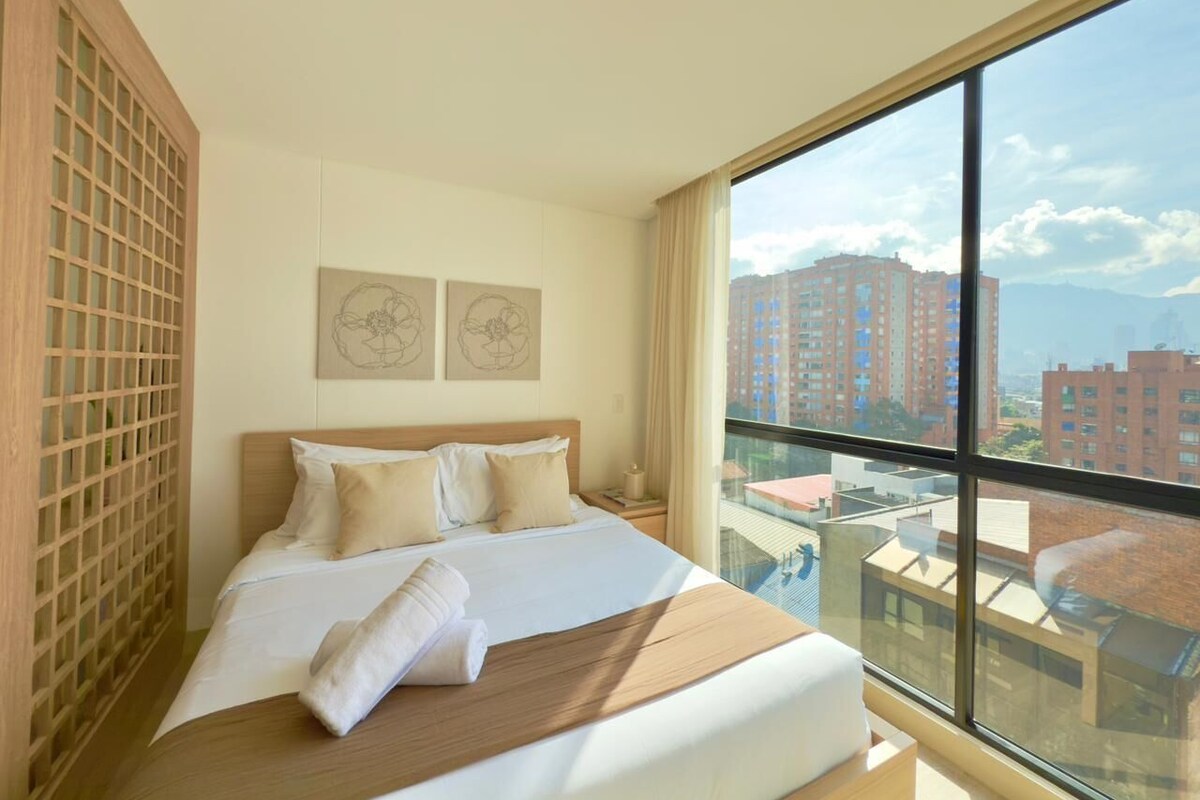 A hotel room with a large bed, a view of tall buildings, and artwork on the wall.