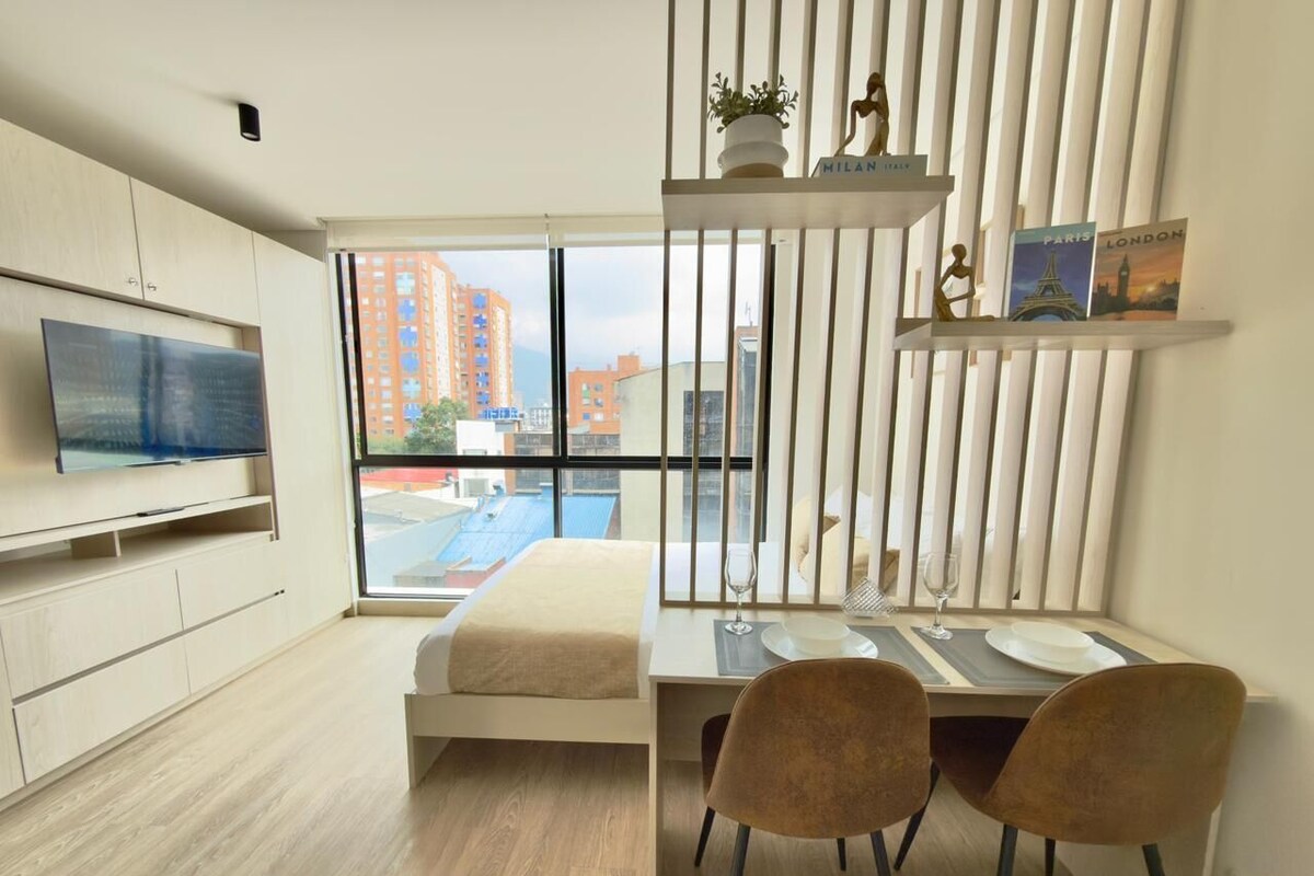 A modern hotel room with a bed, a desk with chairs, a TV, and a city view through large windows.