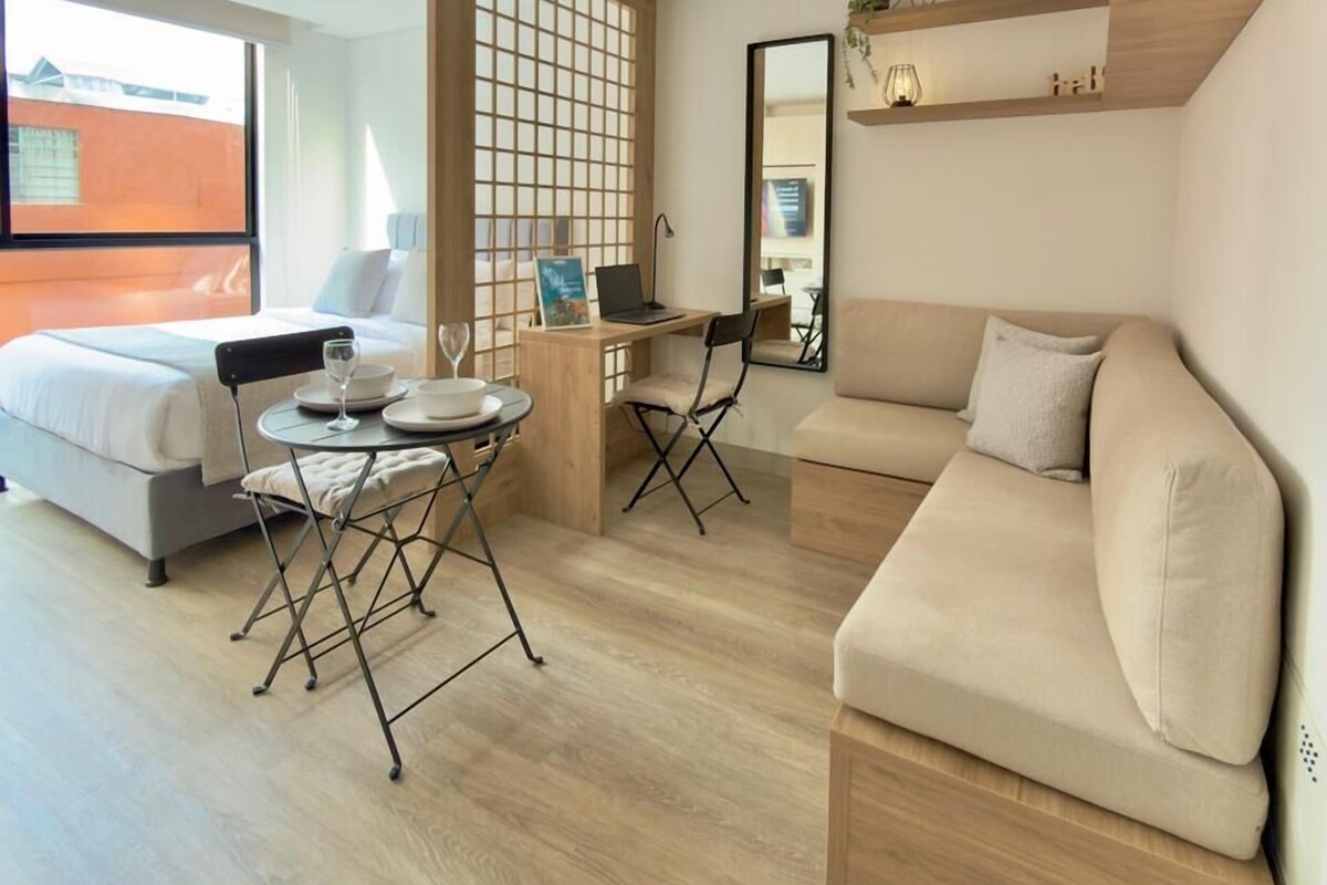 A modern, well-lit room with a bed, a sofa, a small dining table, and a desk. There is a large window providing natural light and a view of the outside.