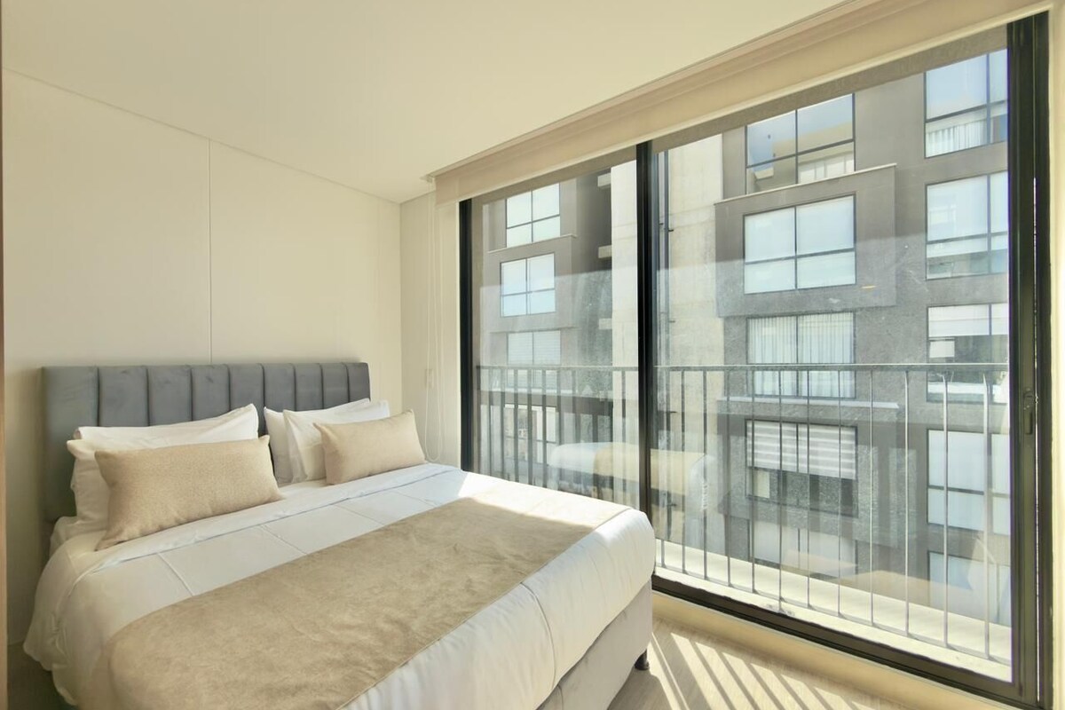 A modern bedroom with a large bed, a grey headboard, and a view of a cityscape through floor-to-ceiling windows.