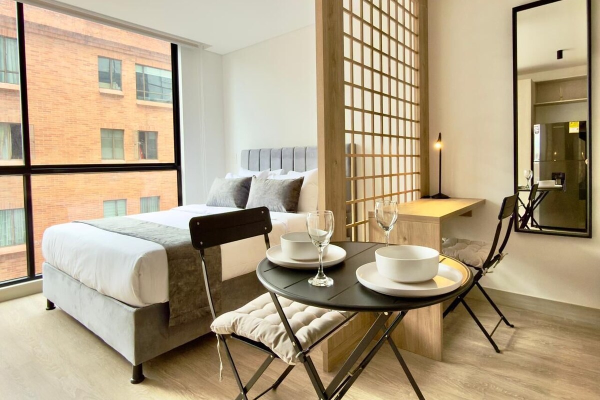 A modern hotel room with a large bed, a dining table with plates and glasses, a wooden partition, and a large window offering a view of a brick building.