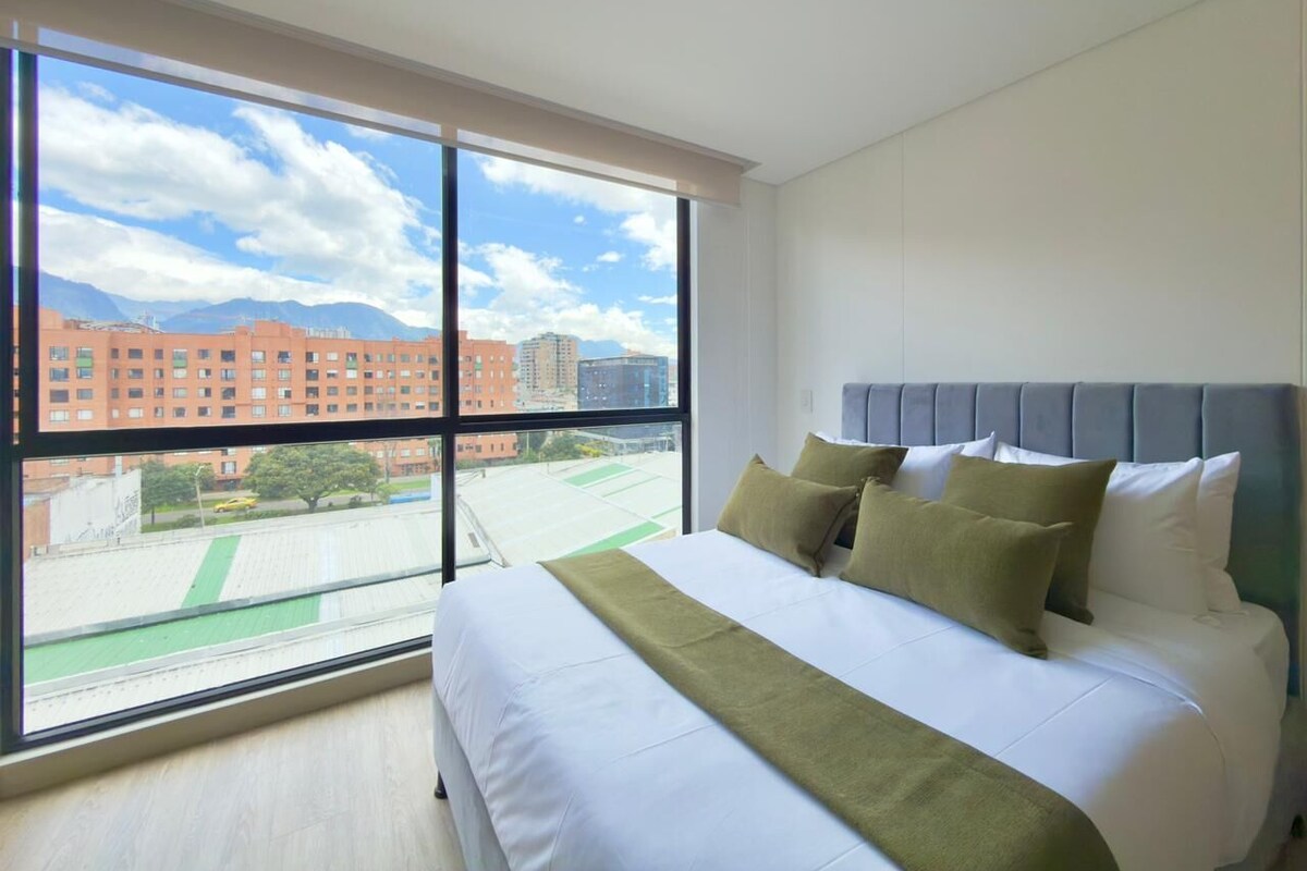 A hotel room with a large window, a bed with white bedding and green throw pillows, and a grey headboard.