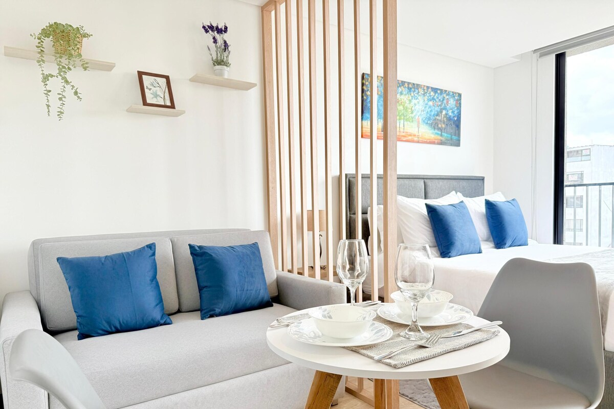 A modern hotel room with a grey sofa, a round dining table with chairs, a bed with blue pillows, and a cityscape view through the window.