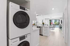 A modern laundry room with stacked washing machines and a kitchen area with white cabinetry and a view of the outdoors. - C-117 - Luxury Park Shore Resort Condo with Pool (Naples)