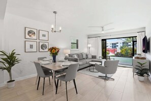 A modern living room with a dining area, a grey sofa, a ceiling fan, and a large window with a view of buildings and greenery. - C-117 - Luxury Park Shore Resort Condo with Pool (Naples)