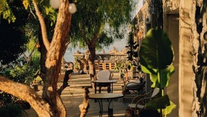 An outdoor patio area with stone flooring, metal tables, and chairs, surrounded by greenery and stone walls. - Maison du monde (Costa Adeje)