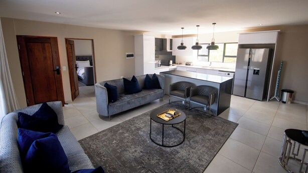 Un salon moderne avec un canapé gris, une table basse et un coin cuisine équipé d’un réfrigérateur, d’une cuisinière et de tabourets de bar.