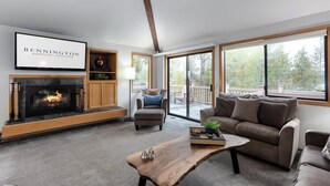 A modern living room with a fireplace, a sofa, a coffee table, and a large window offering a view of the outdoors. - WHEL28 | 4BR • 3.5BA • A/C • SHARC Passes • Bikes (Sunriver)