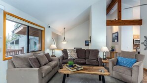 A modern living room with two sofas, a coffee table, and a dining area visible through a large window. - WHEL28 | 4BR • 3.5BA • A/C • SHARC Passes • Bikes (Sunriver)
