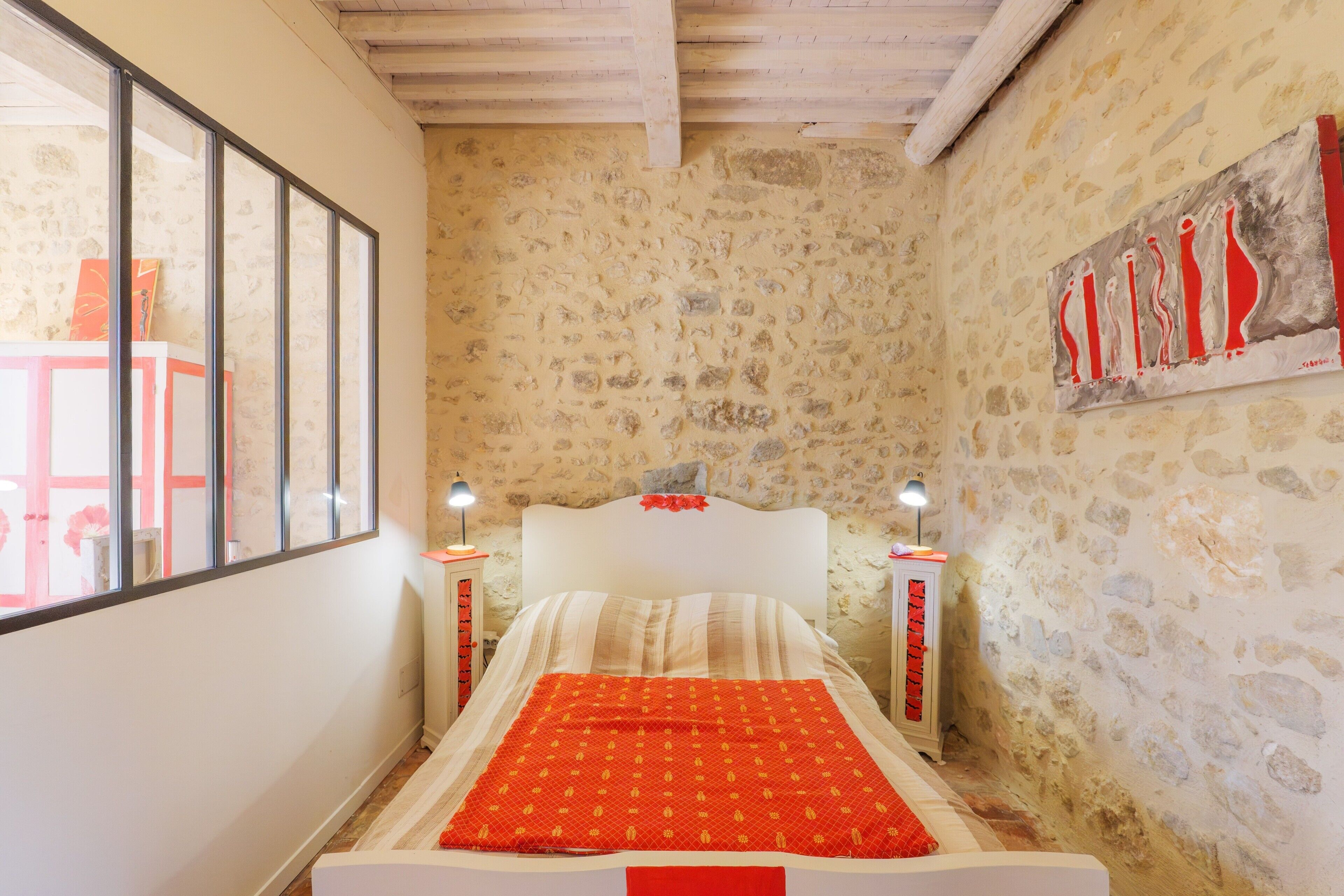 Een slaapkamer met een stenen muur, een bed met een rood-witte dekbedovertrek, twee bedlampen en een raam met uitzicht op een rode stoel en tafel.