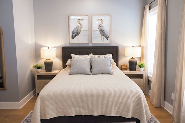 Un dormitorio ordenado con una cama grande, dos mesitas de noche con lámparas, un cabecero y dos cuadros enmarcados.