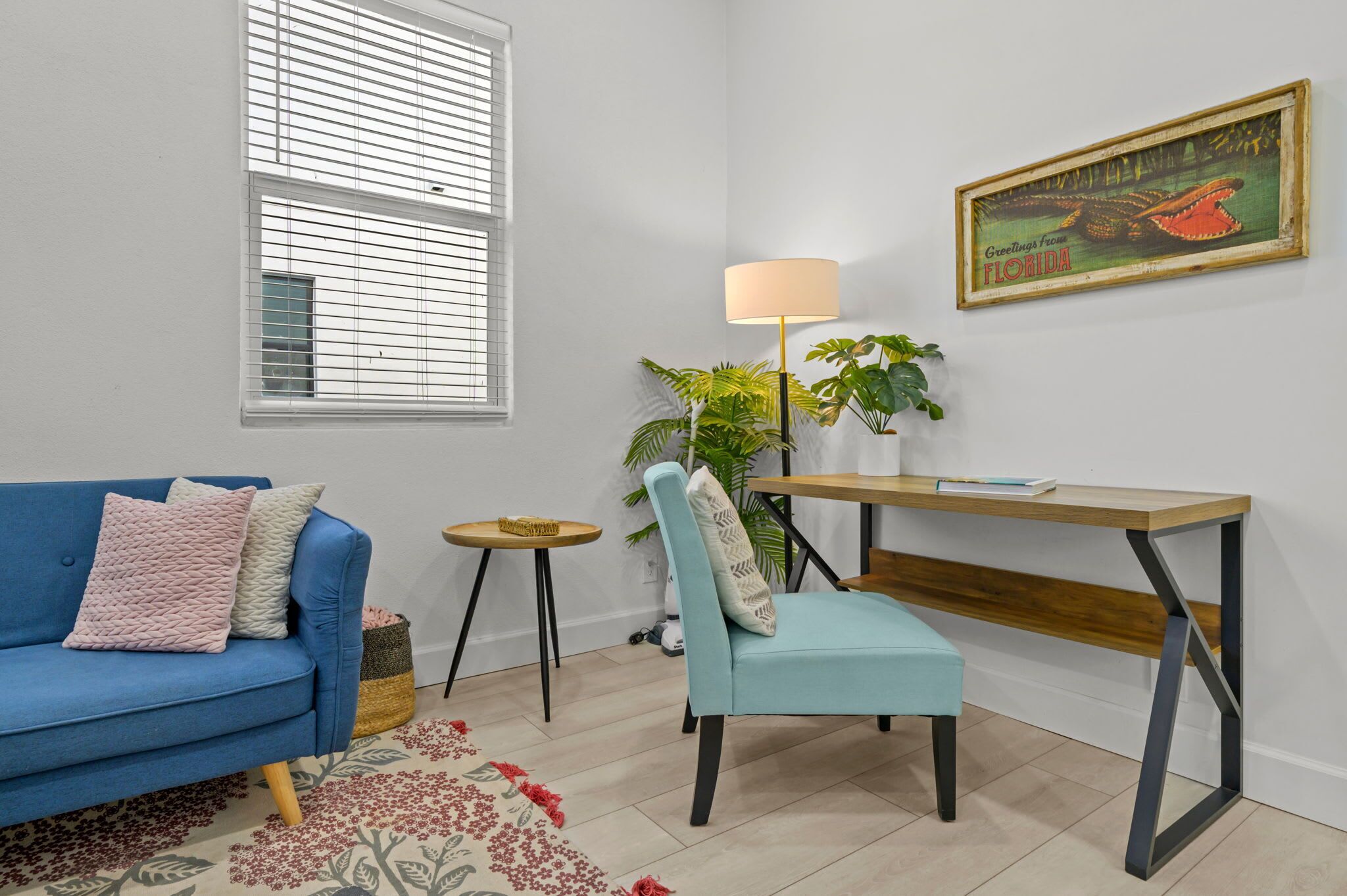 A room with a blue sofa, a light blue chair, a wooden desk, a lamp, a plant, and a framed picture on the wall.