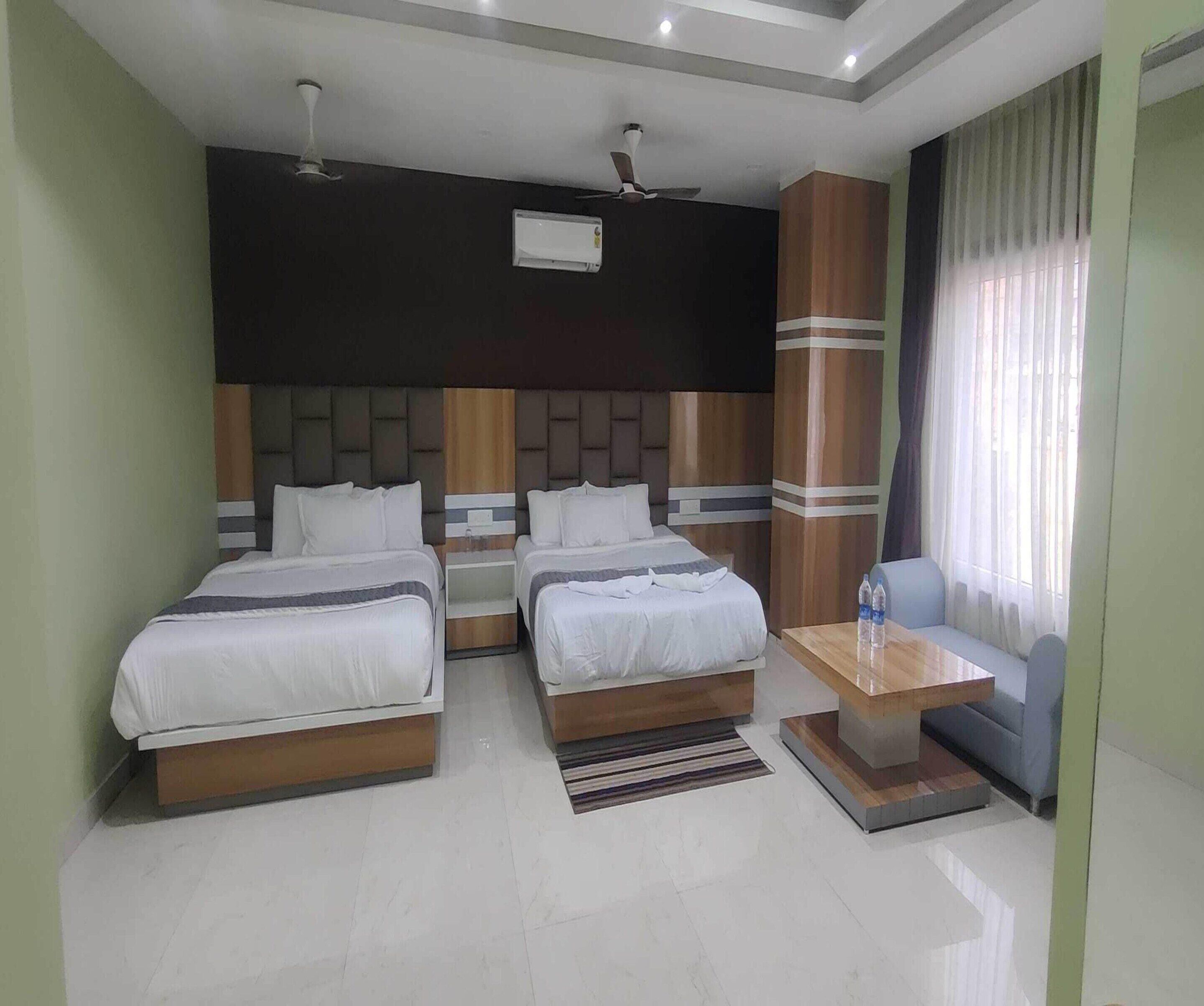 A hotel room with two beds, a small table, a sofa, and a large window with curtains.