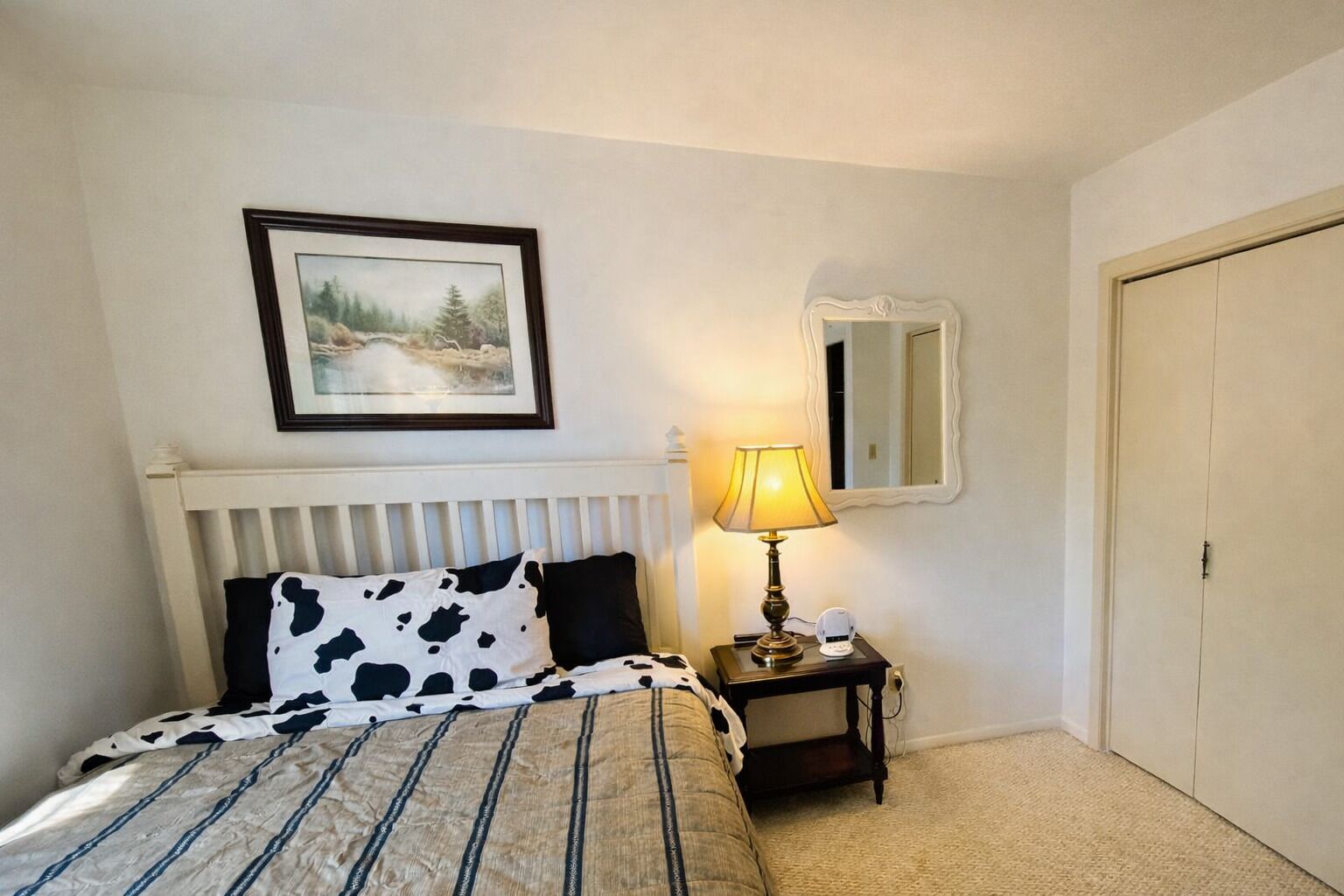 A bedroom with a bed, a bedside table with a lamp, a framed picture on the wall, and a door.