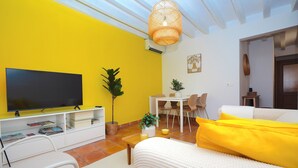 Una habitación luminosa de color amarillo con un televisor de pantalla plana, una mesa de comedor, sillas, un sofá blanco y una cama con almohadones amarillos.