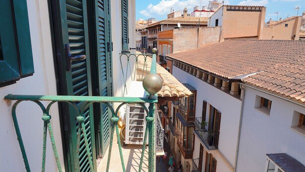 Una calle estrecha flanqueada por edificios, balcones con barandillas de metal y un cielo azul despejado.