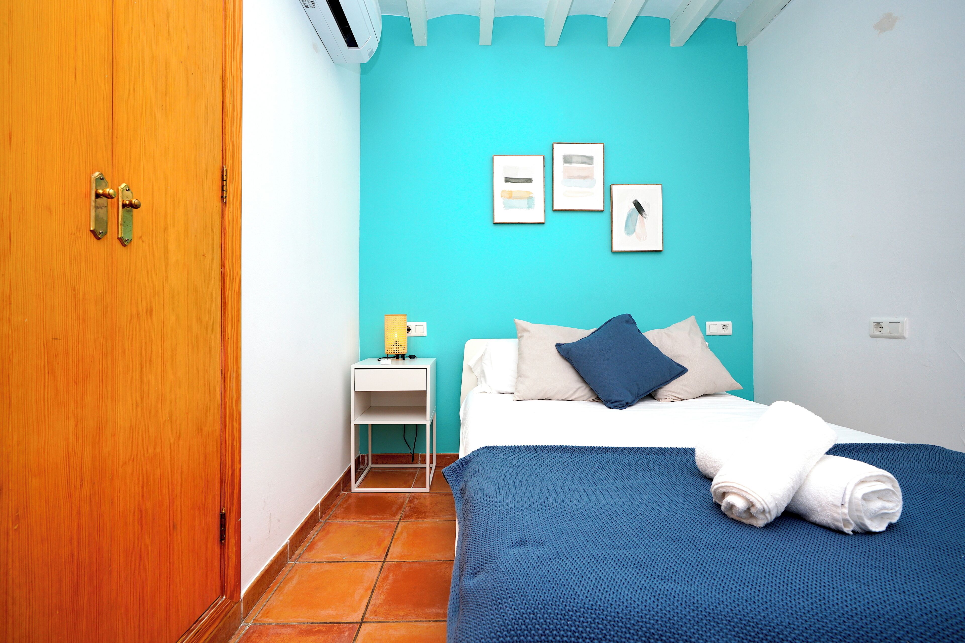 A room with a wooden door, a bed with a blue blanket and white pillows, a white bedside table with a lamp, and a wall with three framed pictures.